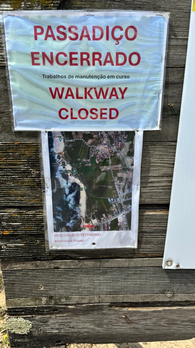 A sign on a wooden surface indicates that a walkway is closed, with text in both Portuguese and English. Below the text, there is a map