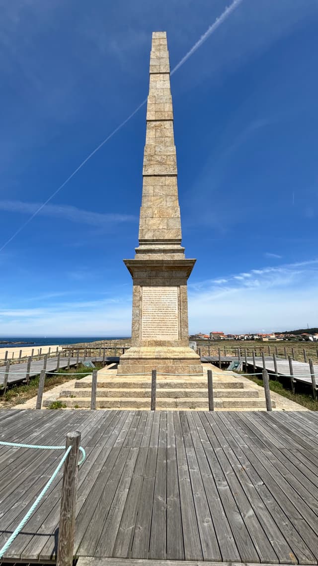 A tall stone obelisk stands on a wooden platform, surrounded by a railing, under a clear blue sky