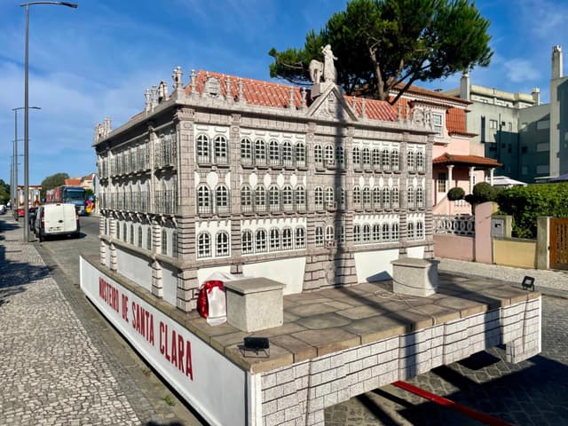 A detailed architectural model of a historic building is displayed on a platform outdoors, surrounded by a street and residential area
