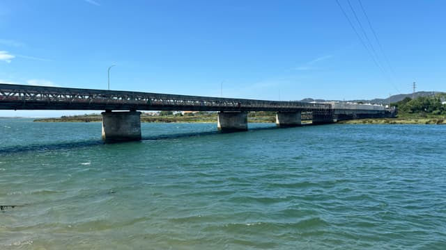 A long bridge spans across a body of water under a clear blue sky, with greenery visible in the background