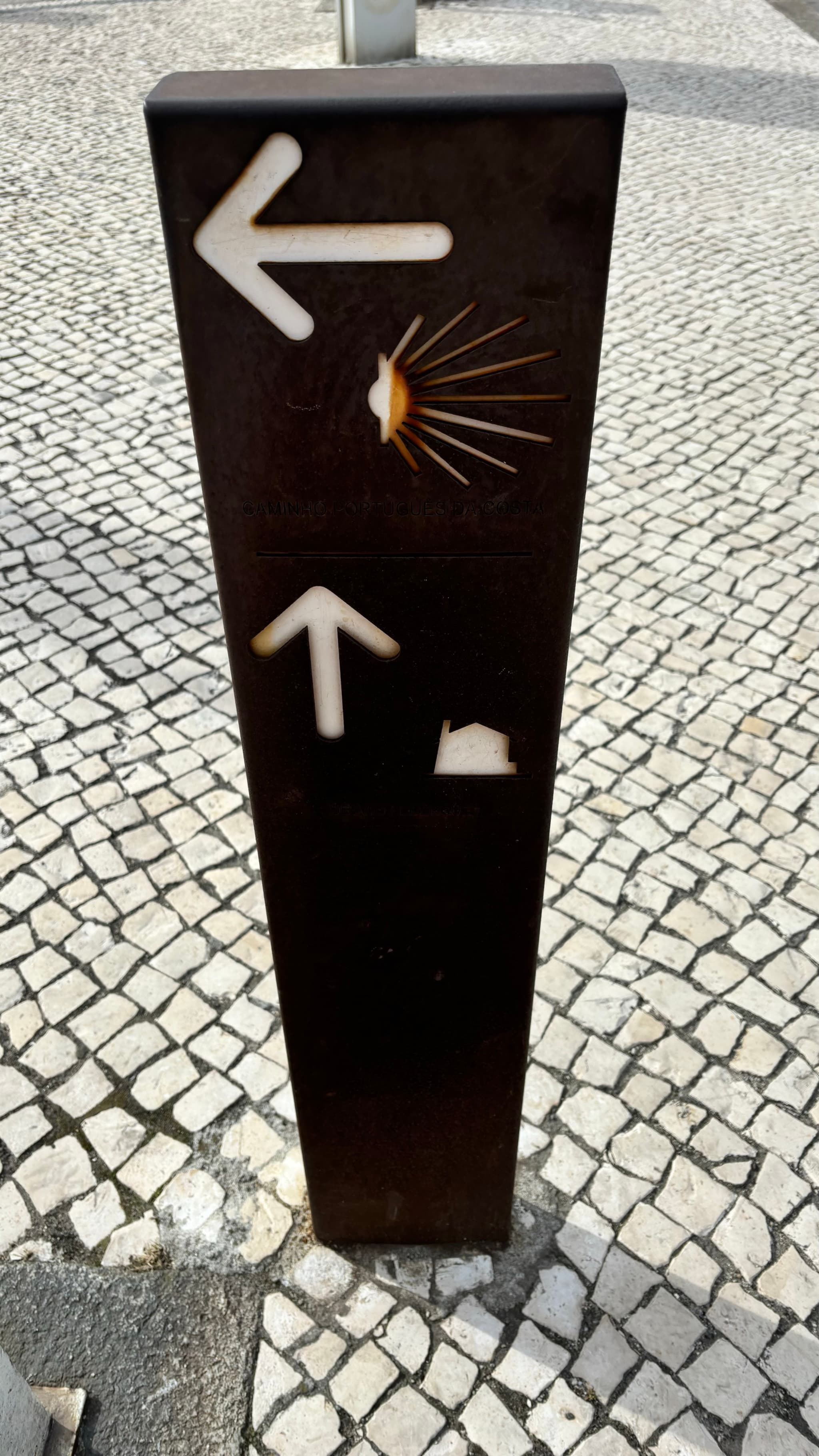 A signpost with two directional arrows, one pointing left with a shell symbol and one pointing up with a house symbol, set on a cobblestone pavement