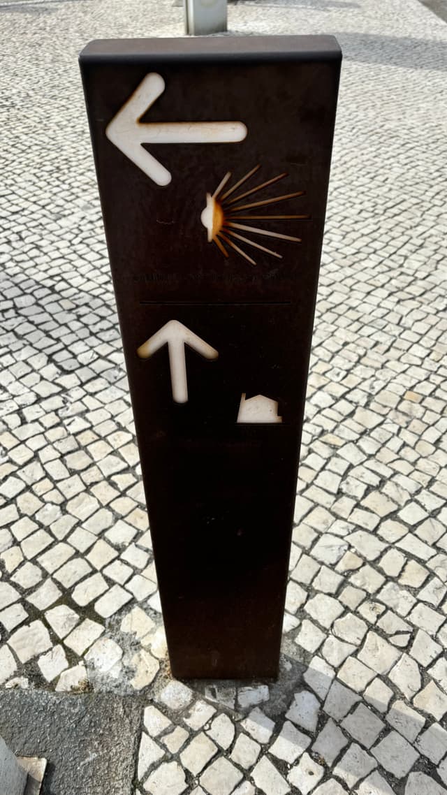 A signpost with two directional arrows, one pointing left with a shell symbol and one pointing up with a house symbol, set on a cobblestone pavement