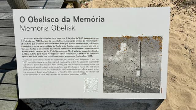 A sign titled O Obelisco da Memória with text in Portuguese and English, accompanied by a historical photograph of the Memória Obelisk. The sign is placed outdoors on a sandy surface