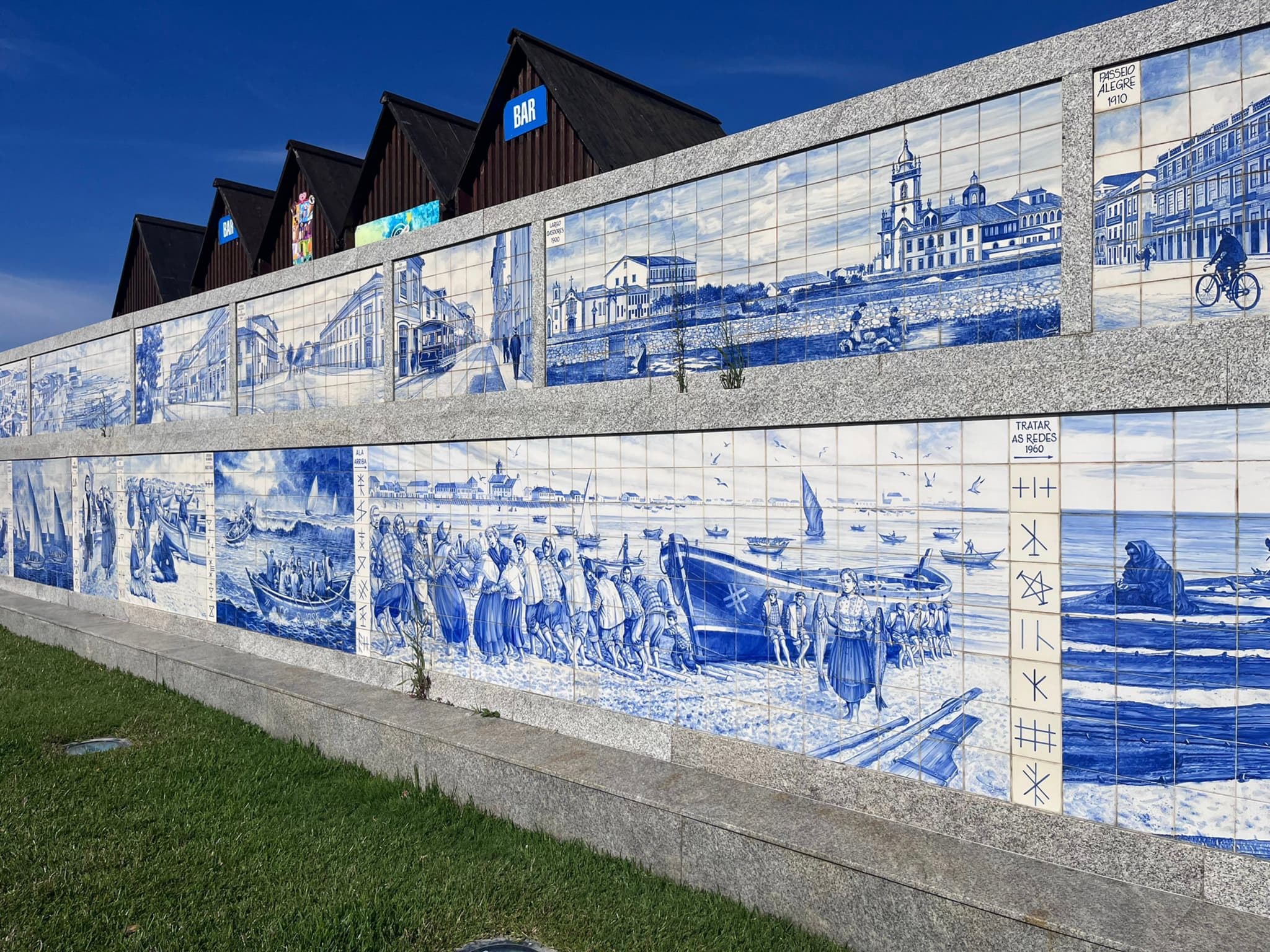 A wall adorned with blue and white tiles depicting various scenes, set against a backdrop of buildings with triangular roofs and a clear blue sky