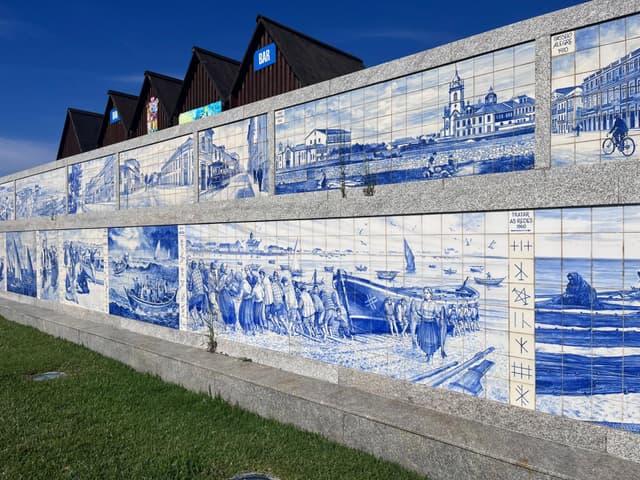 A wall adorned with blue and white tiles depicting various scenes, set against a backdrop of buildings with triangular roofs and a clear blue sky