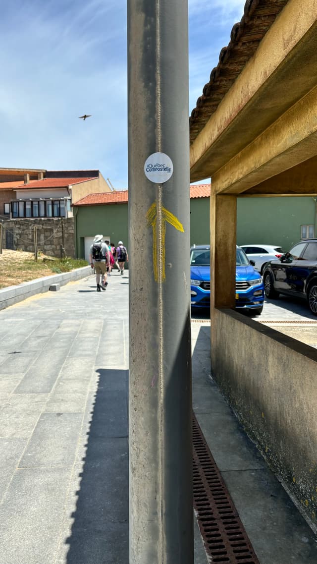 A street scene with a metal pole featuring a yellow arrow and a circular sticker. People are walking in the background, and cars are parked on the right. A bird is flying in the sky