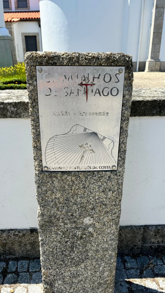 A stone marker with a metal plaque indicating the Caminhos de Santiago route, featuring a scallop shell design and directional lines