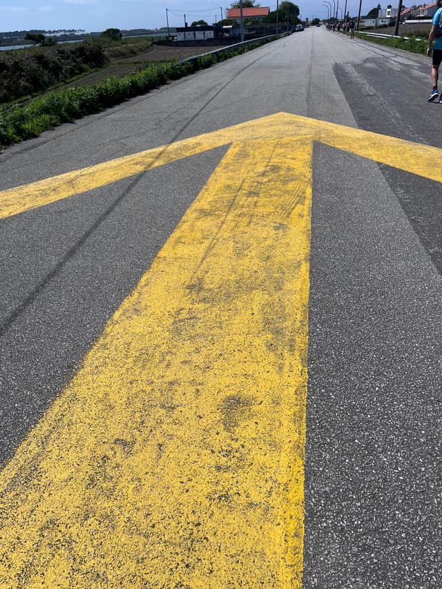 A large yellow arrow painted on a road, pointing straight ahead, with a clear sky and distant buildings in the background