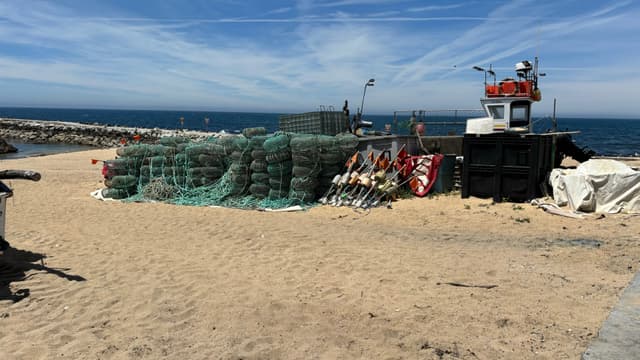 A sandy beach with stacked fishing nets, buoys, and a small boat near the shoreline under a blue sky