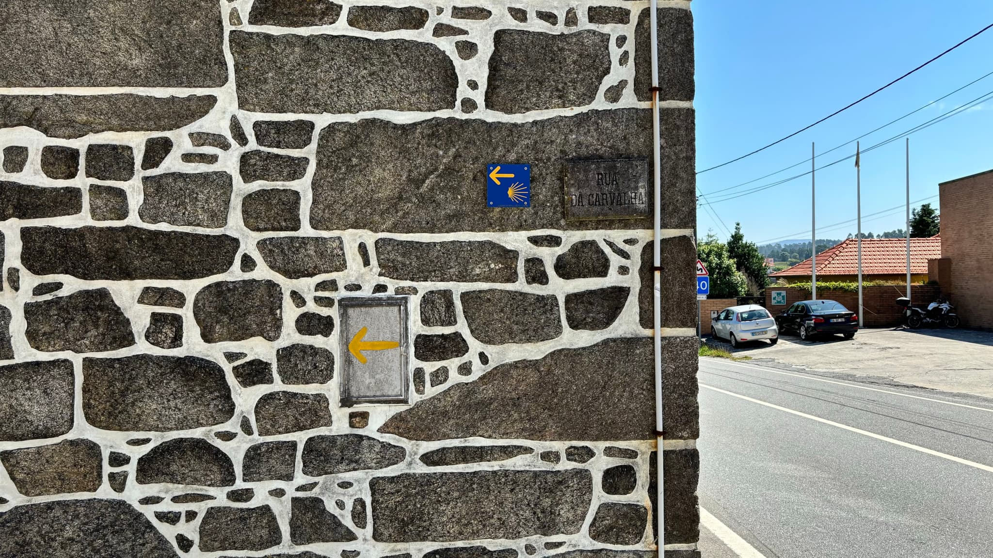 A stone wall with a blue Camino de Santiago sign and a yellow arrow. A road and parked cars are visible in the background under a clear blue sky