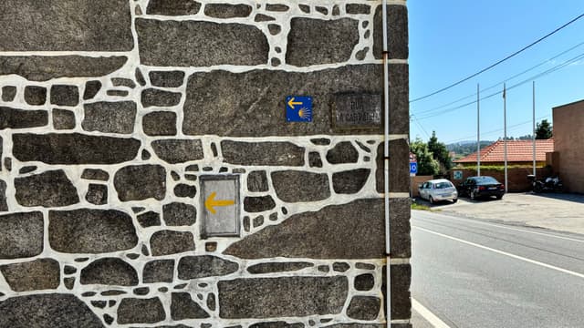 A stone wall with a blue Camino de Santiago sign and a yellow arrow. A road and parked cars are visible in the background under a clear blue sky