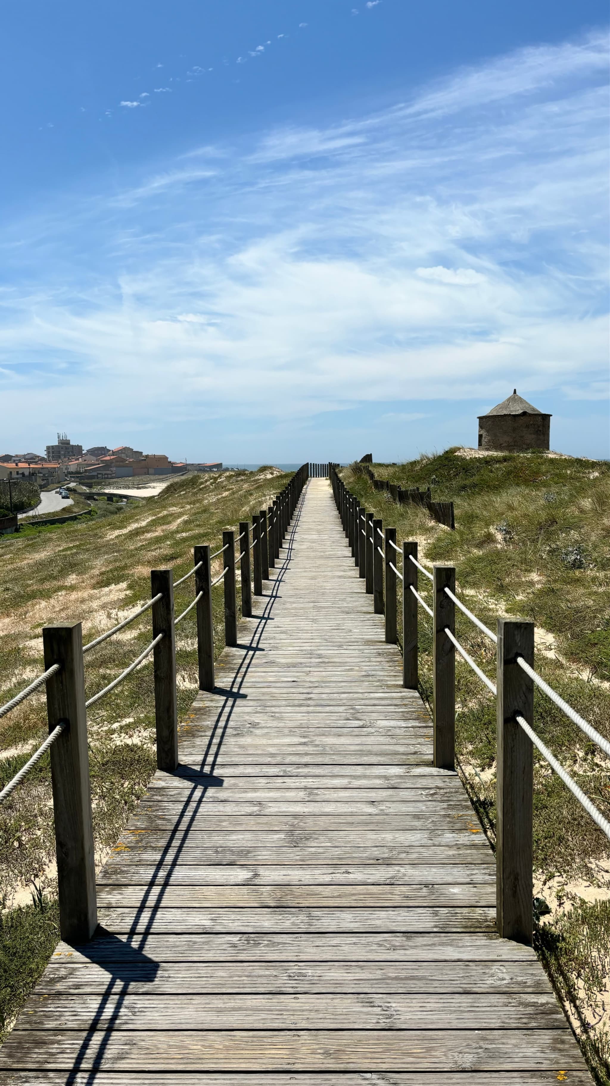 A wooden boardwalk stretches through grassy dunes under a blue sky, with a small stone structure on the right and distant buildings on the left
