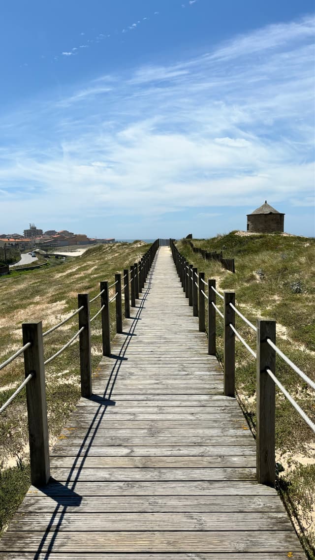 A wooden boardwalk stretches through grassy dunes under a blue sky, with a small stone structure on the right and distant buildings on the left