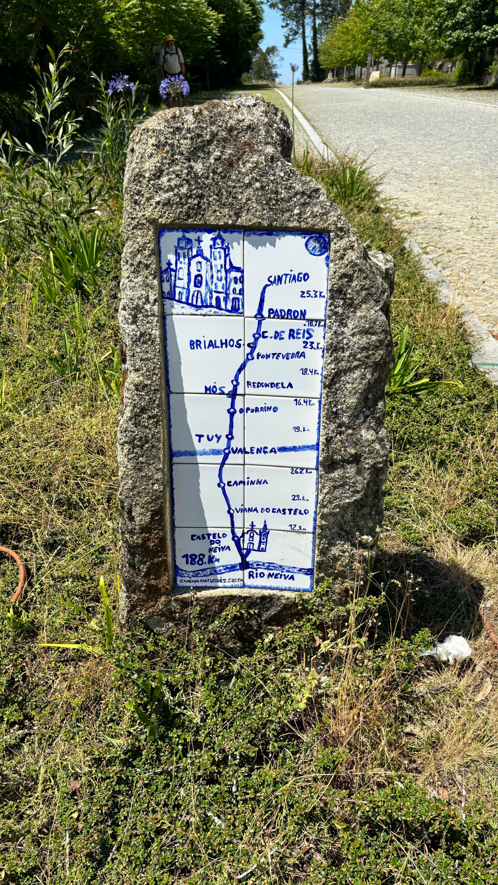 A stone marker with a blue and white ceramic tile displays a map and distances to various locations, set beside a road with greenery around