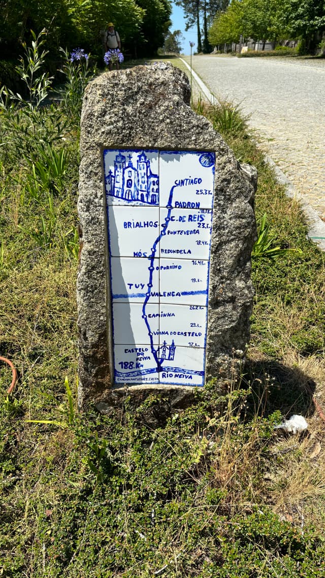 A stone marker with a blue and white ceramic tile displays a map and distances to various locations, set beside a road with greenery around