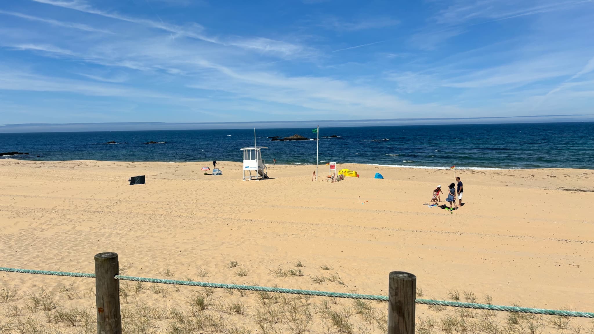 A sandy beach with a lifeguard tower, a few people, and colorful umbrellas near the shoreline, under a clear blue sky