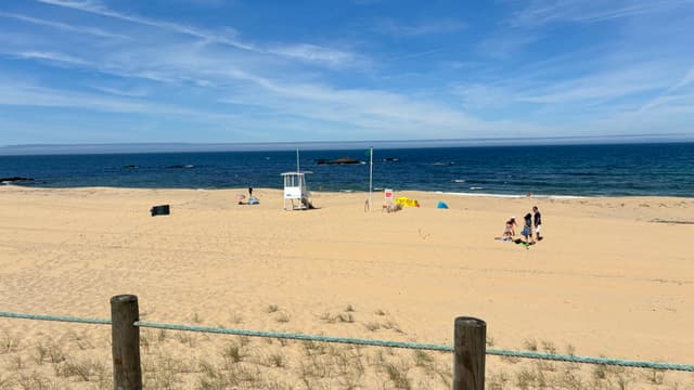 A sandy beach with a lifeguard tower, a few people, and colorful umbrellas near the shoreline, under a clear blue sky