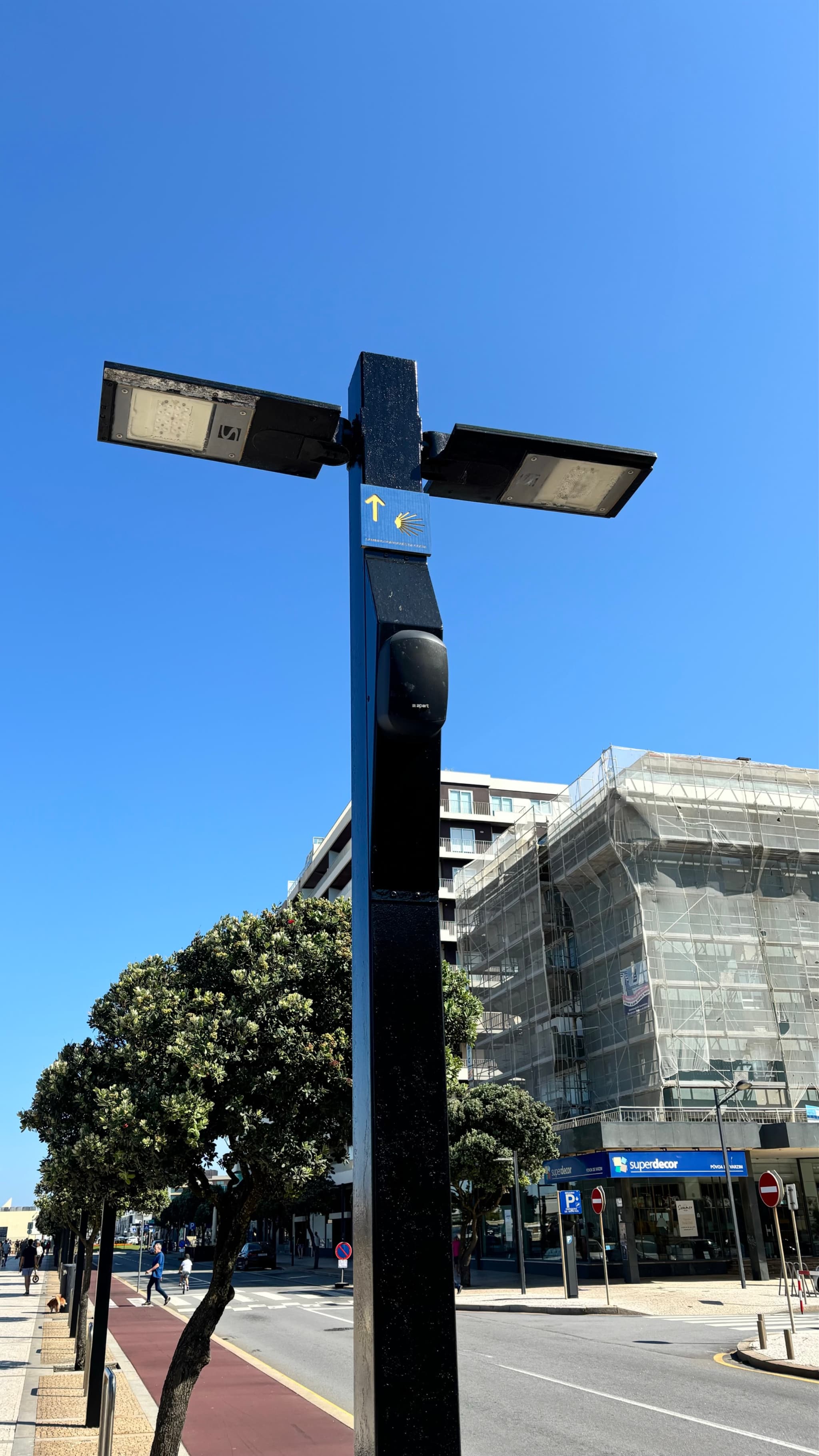 A streetlight with two lamps on a pole, set against a clear blue sky Nearby, a building is under construction with scaffolding, and there are trees and a road in the background