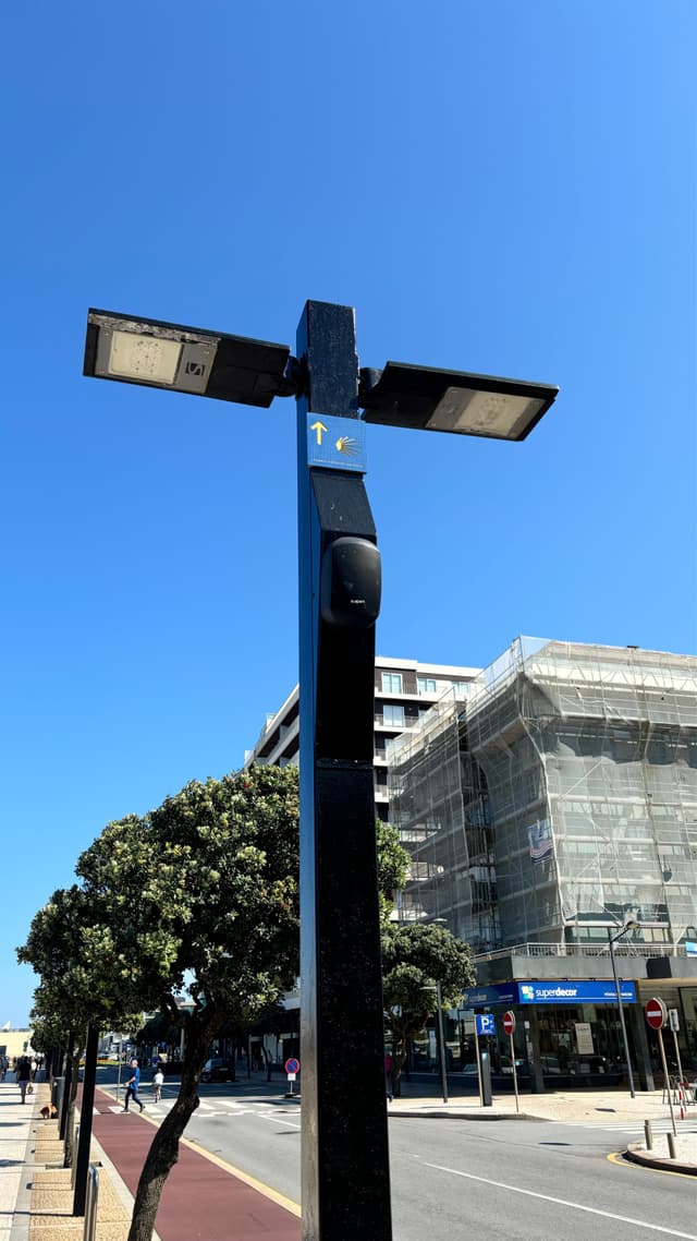 A streetlight with two lamps on a pole, set against a clear blue sky Nearby, a building is under construction with scaffolding, and there are trees and a road in the background