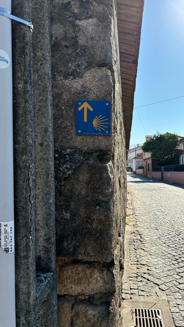 A stone wall with a blue sign featuring a yellow arrow and a scallop shell, indicating a direction along a cobblestone street