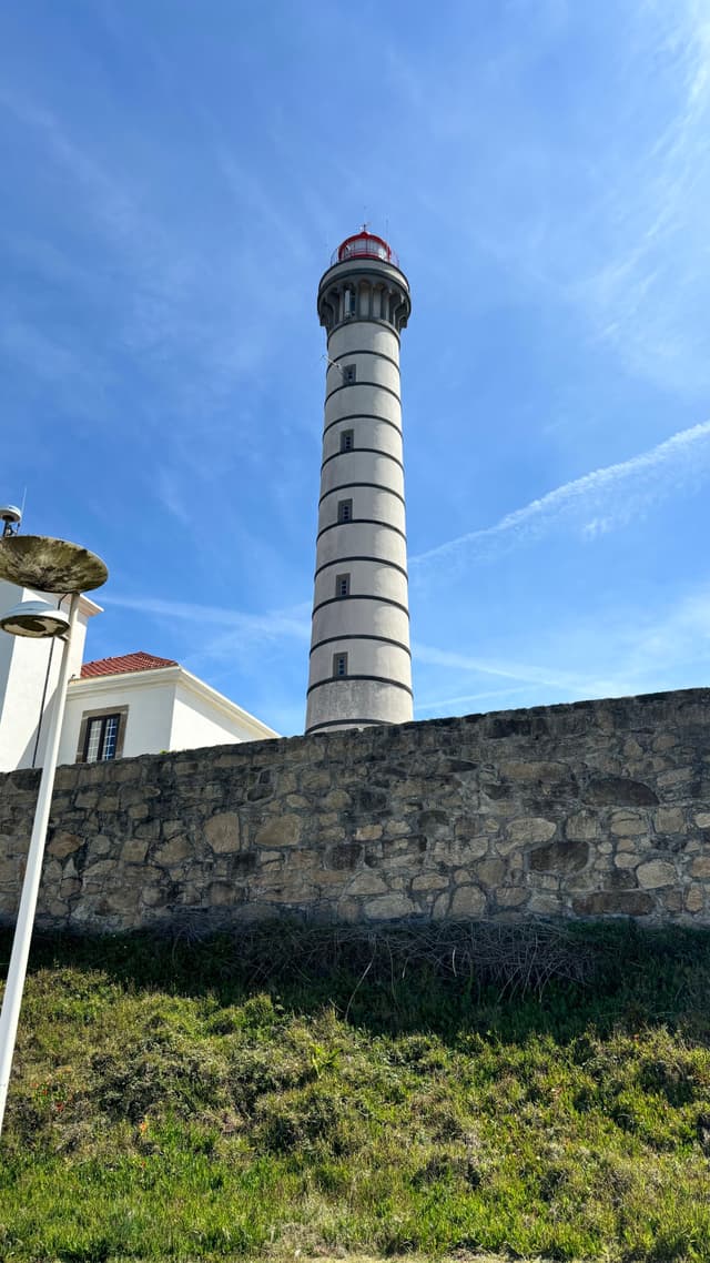 A tall, cylindrical lighthouse with horizontal stripes stands behind a stone wall, under a clear blue sky