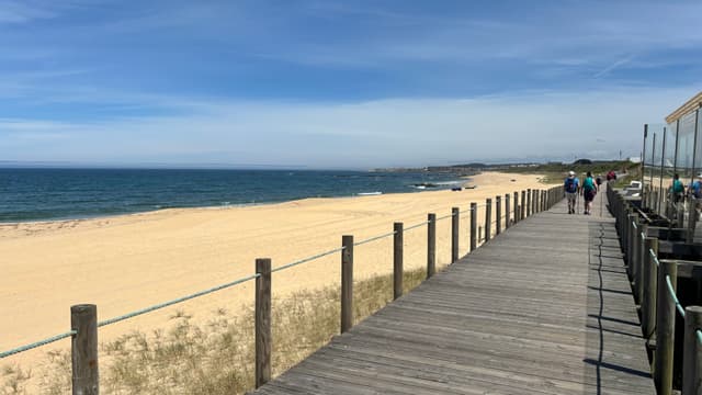 A wooden boardwalk runs alongside a sandy beach with a clear blue sky and ocean. People are walking along the boardwalk, and there are some structures on the right side