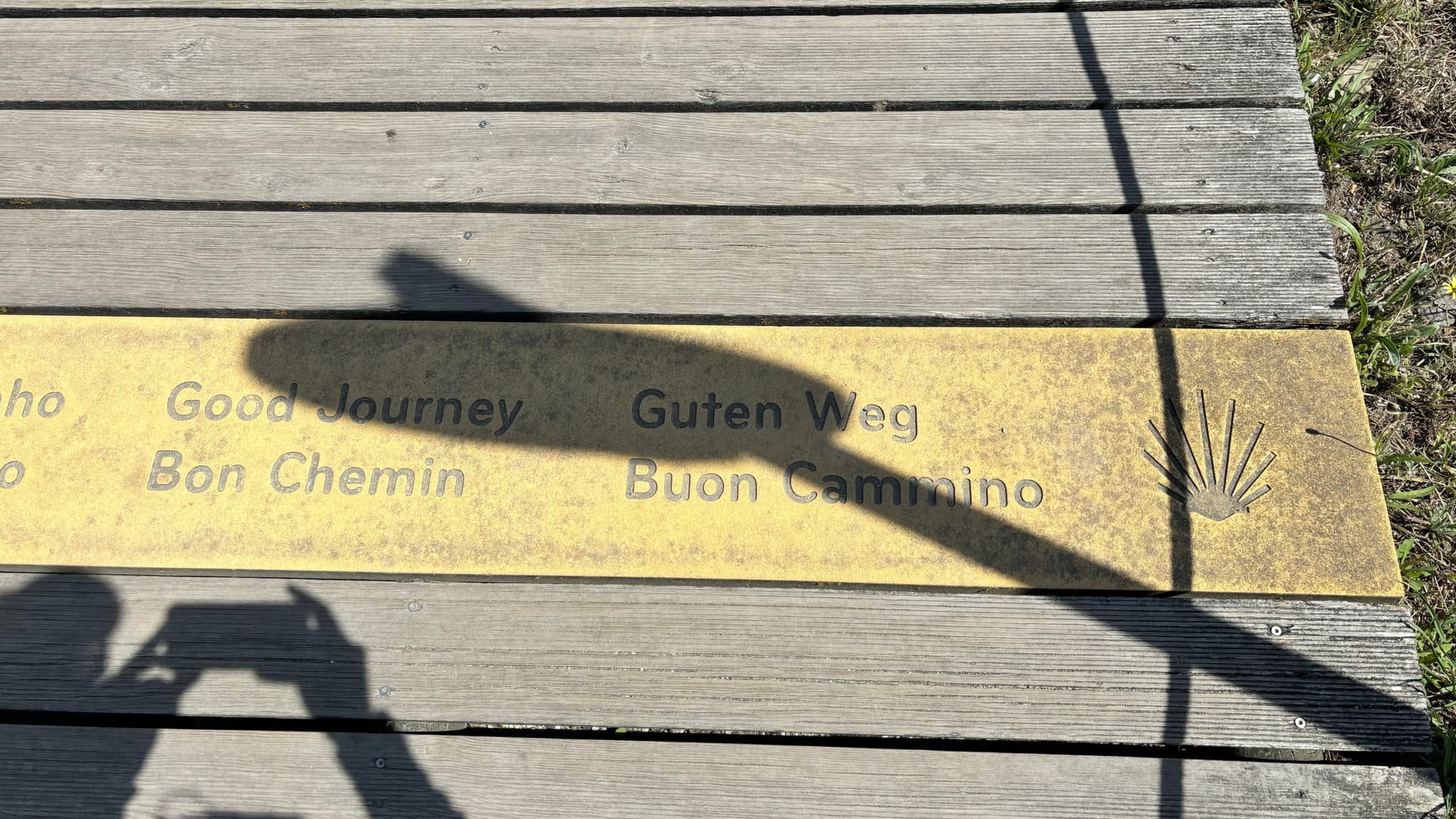 A wooden walkway with a metal plaque displaying the words Good Journey in multiple languages, including Guten Weg and Buen Camino, alongside a scallop shell symbol. A shadow of a person taking a photo is visible