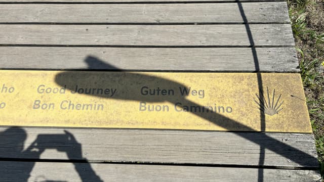 A wooden walkway with a metal plaque displaying the words Good Journey in multiple languages, including Guten Weg and Buen Camino, alongside a scallop shell symbol. A shadow of a person taking a photo is visible