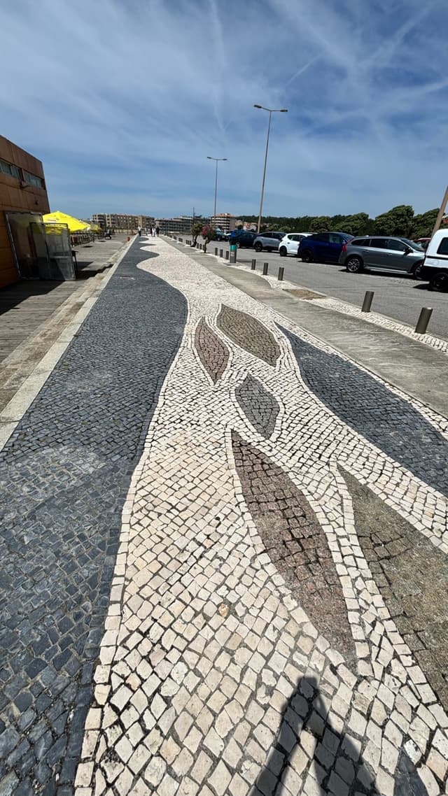 A patterned stone walkway with abstract designs runs alongside a street with parked cars and a clear blue sky above