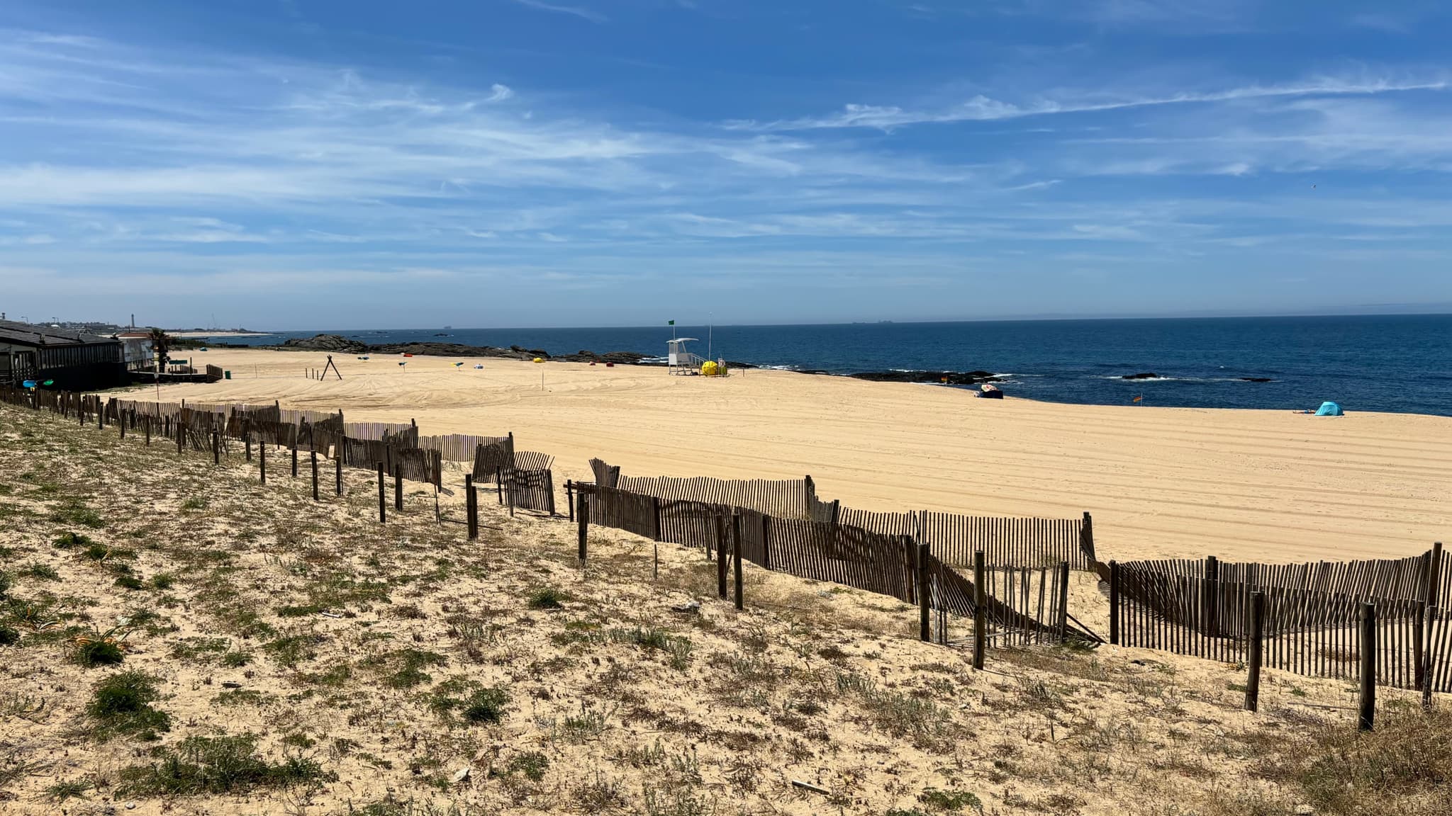 A sandy beach with a wooden fence, sparse vegetation, and a clear blue sky. The ocean is visible in the background