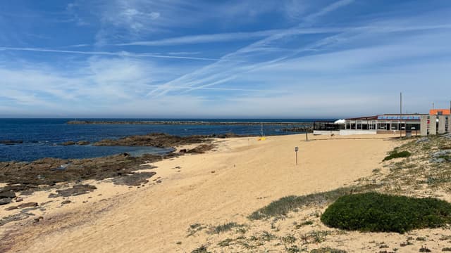 A sandy beach with scattered rocks along the shoreline, a clear blue sky with wispy clouds, and a building near the water's edge