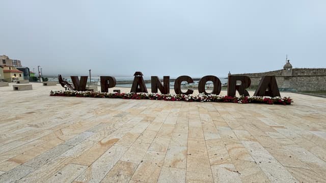 Large letters spelling V.P. ÂNCORA on a paved area, with a cloudy sky and buildings in the background