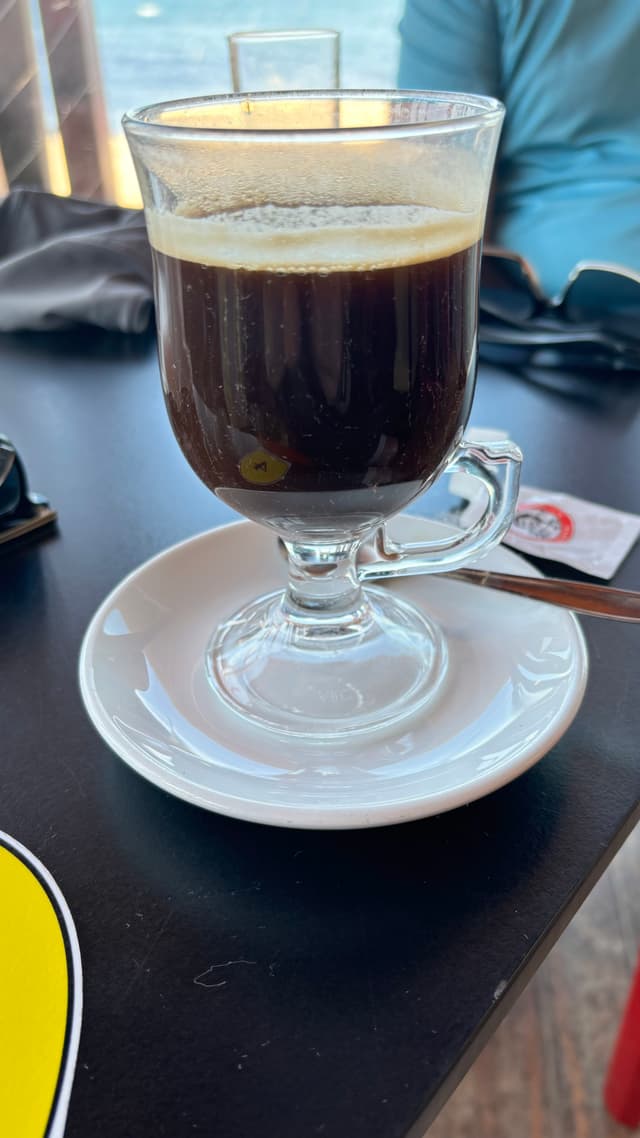 A glass mug filled with black coffee sits on a white saucer on a dark table, with a spoon and a sugar packet nearby