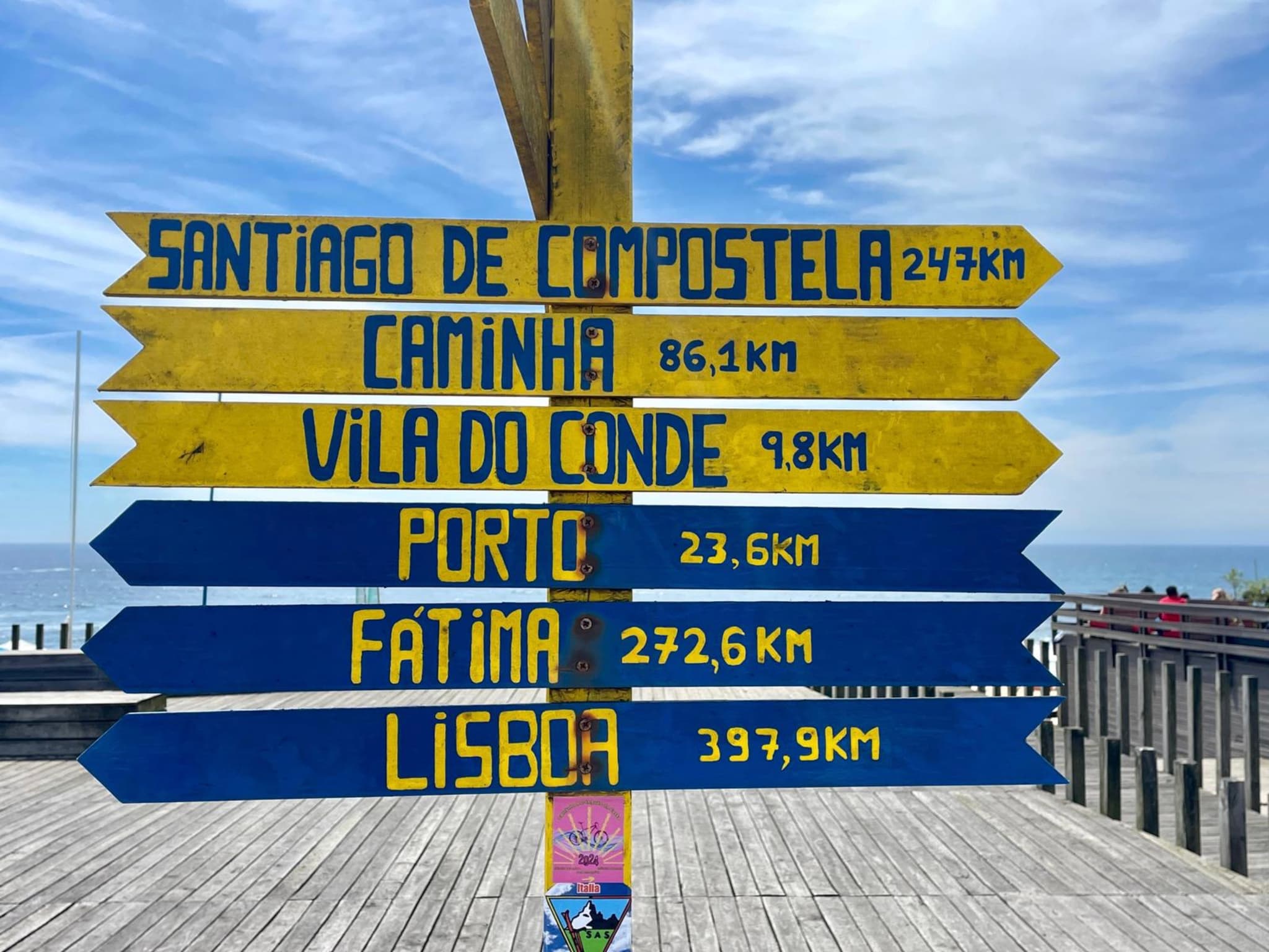 A colorful signpost with directional arrows points to various locations, including Santiago de Compostela, Caminha, Vila do Conde, Porto, Fátima, and Lisboa, with distances in kilometers. The background features a wooden deck and a blue sky