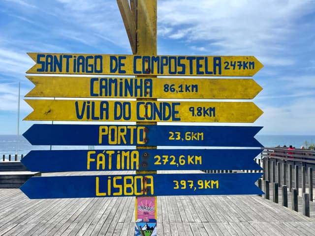 A colorful signpost with directional arrows points to various locations, including Santiago de Compostela, Caminha, Vila do Conde, Porto, Fátima, and Lisboa, with distances in kilometers. The background features a wooden deck and a blue sky