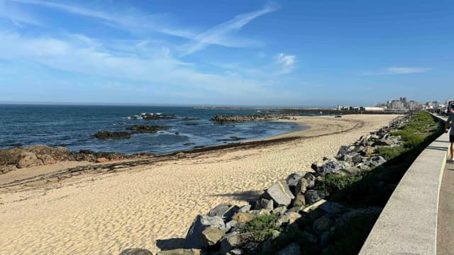 A sandy beach with scattered rocks along the shoreline, bordered by a stone wall on the right, under a clear blue sky.