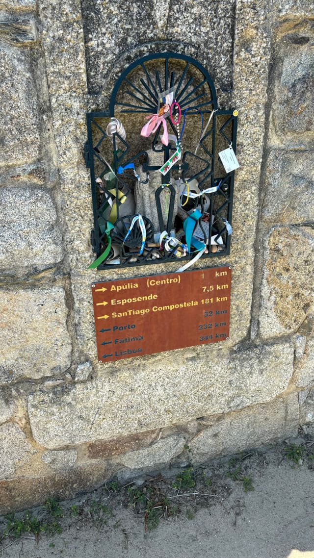 A stone wall features a small shrine with colorful ribbons and trinkets attached. Below, a plaque displays distances to various locations, including Santiago de Compostela