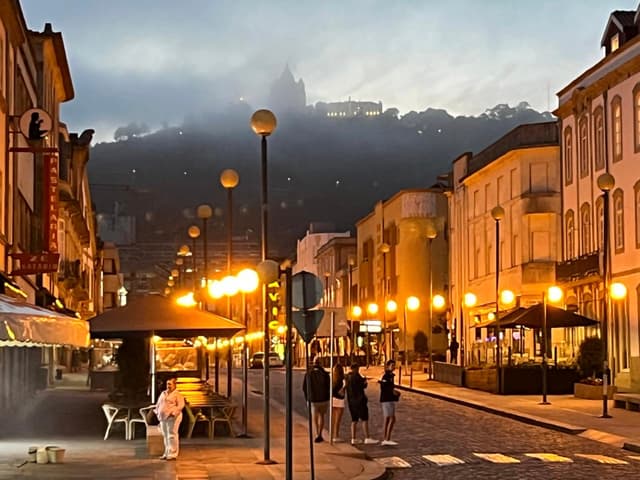 A cobblestone street lined with illuminated buildings and streetlights at dusk, with people walking and a misty hill in the background