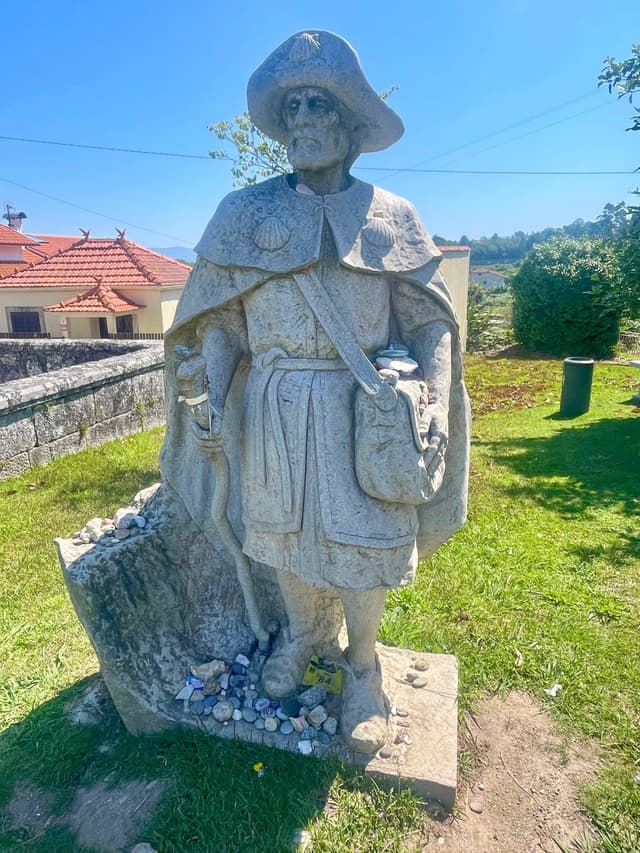 A stone statue of a historical figure wearing a cloak and hat, holding a staff, stands on a grassy area with small stones at its base