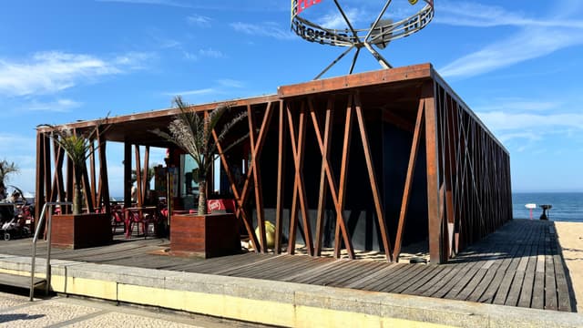 A beachside structure with wooden beams and a patio, featuring outdoor seating and palm plants, set against a clear blue sky and ocean backdrop