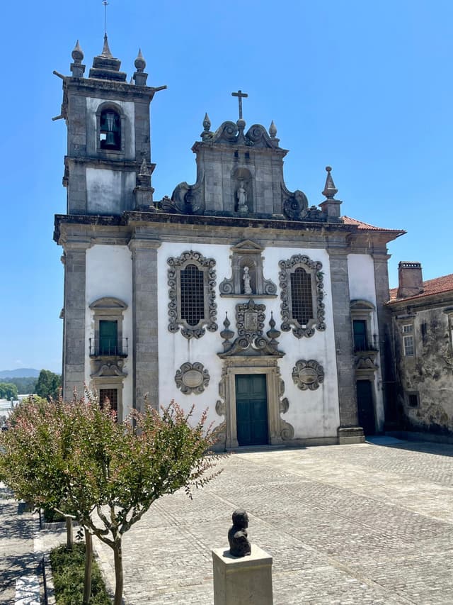 A historic church with ornate architectural details, featuring a bell tower and decorative facade, set against a clear blue sky. A small tree and a sculpture are in the foreground