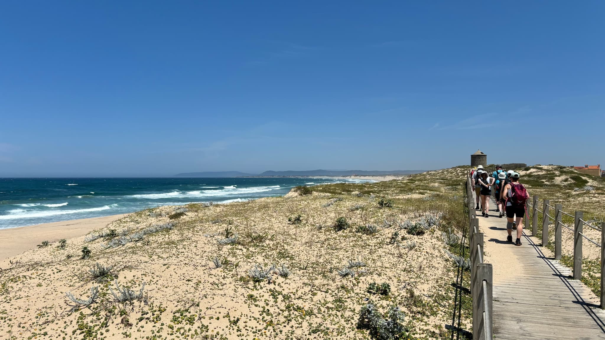 A coastal landscape with a sandy beach and ocean waves on the left, a wooden boardwalk on the right, and people walking along it under a clear blue sky