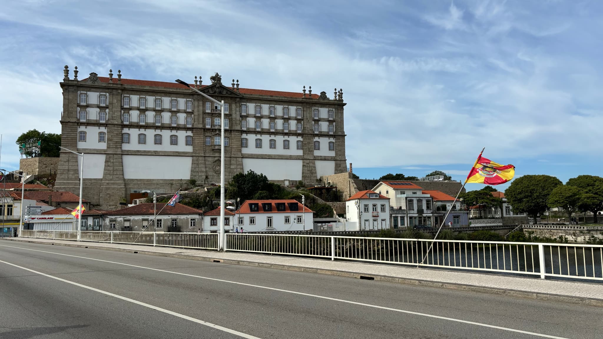 A large, historic building with a red-tiled roof stands near a river, with a bridge in the foreground and a colorful flag waving on the right