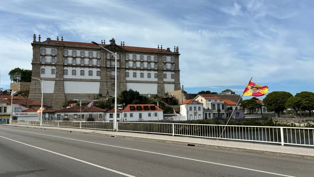A large, historic building with a red-tiled roof stands near a river, with a bridge in the foreground and a colorful flag waving on the right